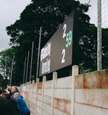 A large digital scoreboard at an outdoor sports venue displays a score of 2-2. Spectators are gathered, observing the event with interest. The scene is enclosed by a tall metal fence and surrounded by lush green trees, indicating a natural setting. Overcast skies add a muted tone to the environment.