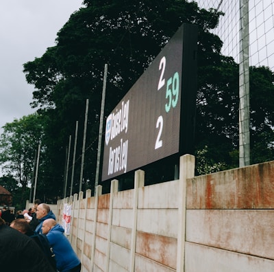 A large digital scoreboard at an outdoor sports venue displays a score of 2-2. Spectators are gathered, observing the event with interest. The scene is enclosed by a tall metal fence and surrounded by lush green trees, indicating a natural setting. Overcast skies add a muted tone to the environment.