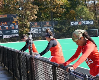 A group of athletes wearing orange uniforms are on the sideline of a sports field, resting against a fence. Behind them, a large digital scoreboard displays team names and scores. The field is bright green, and several banners with sponsor logos and championship wins are visible. The athletes appear to be taking a break, possibly before or during a field hockey game.