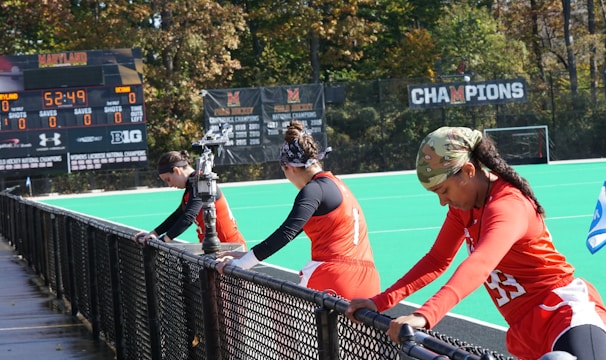 A group of athletes wearing orange uniforms are on the sideline of a sports field, resting against a fence. Behind them, a large digital scoreboard displays team names and scores. The field is bright green, and several banners with sponsor logos and championship wins are visible. The athletes appear to be taking a break, possibly before or during a field hockey game.