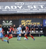 Players engage in a competitive soccer game on a field while others are seated on the bench watching attentively. The action is taking place in front of a wooden structure displaying multiple advertisements, including orthodontics, lacrosse, and field hockey.