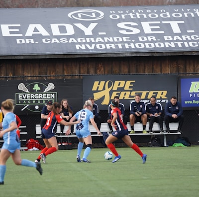 Players engage in a competitive soccer game on a field while others are seated on the bench watching attentively. The action is taking place in front of a wooden structure displaying multiple advertisements, including orthodontics, lacrosse, and field hockey.