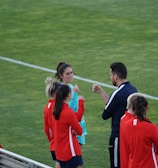A group of people wearing sports attire are gathered on a grassy field. Some individuals are dressed in red jackets, while others are in turquoise and navy blue. A coach appears to be giving instructions. Sports equipment, such as water bottles and a bench with towels, can be seen nearby.