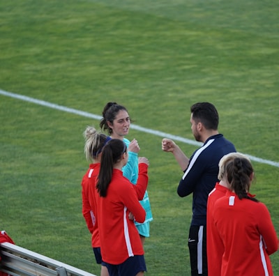 A group of people wearing sports attire are gathered on a grassy field. Some individuals are dressed in red jackets, while others are in turquoise and navy blue. A coach appears to be giving instructions. Sports equipment, such as water bottles and a bench with towels, can be seen nearby.