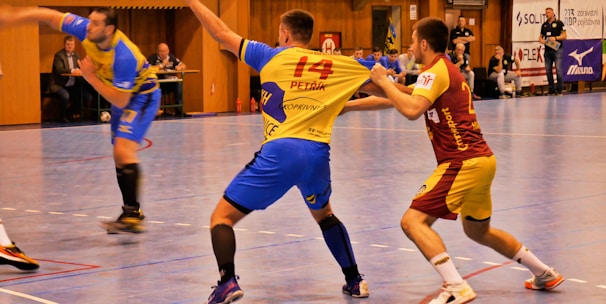 A dynamic indoor sports scene with players in motion on a court. Two athletes in blue and yellow uniforms are engaged in an intense handball game, with one player gripping the other's jersey. The background shows other players, several seated people, and advertisements on the walls.