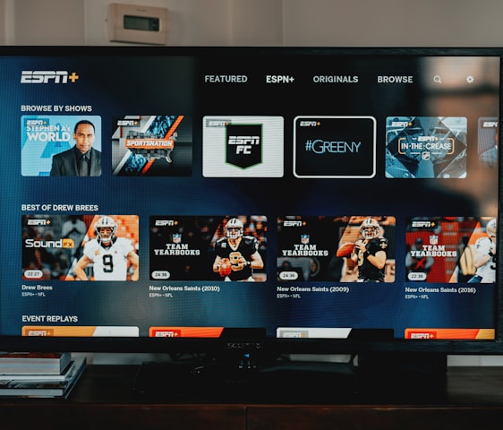 A television screen displaying the ESPN+ streaming interface with various sports-related shows and categories. The screen includes sections such as 'Browse by Shows' and 'Best of Drew Brees' with thumbnails of sports programs featuring athletes in action and analysts.