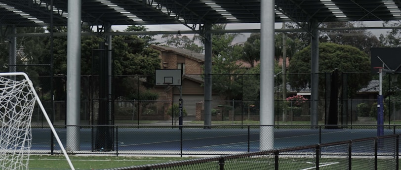 An outdoor sports facility featuring a running track and a soccer goal. The track is red, surrounded by a black metal fence, and the field is covered with artificial green turf. In the background, a basketball court is visible under a covered roof with metal supports. The sky is overcast, adding a moody atmosphere.