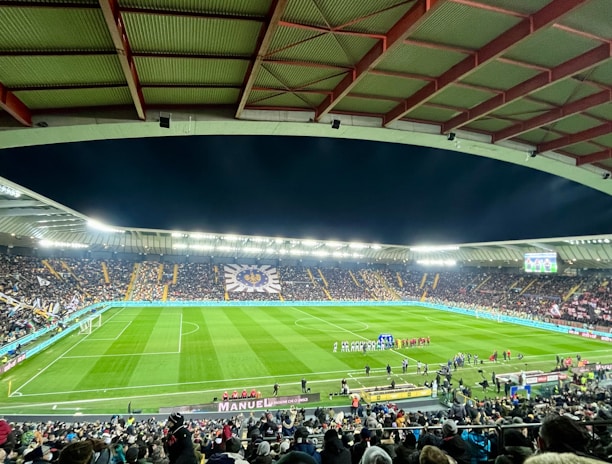 A large football stadium filled with spectators. The field is brightly lit, and two teams are positioned on the pitch. The stadium is covered, with a visible roof structure and stands filled with fans. Scoreboards are visible displaying game information.