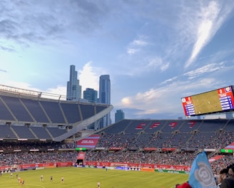 A sports stadium filled with spectators watching a soccer match. Players are on the field in action, and a large scoreboard displays information about the game. The stadium has blue seating and is surrounded by tall city buildings visible in the background against a partly cloudy sky. Fans are dressed casually, some waving flags and wearing team colors.