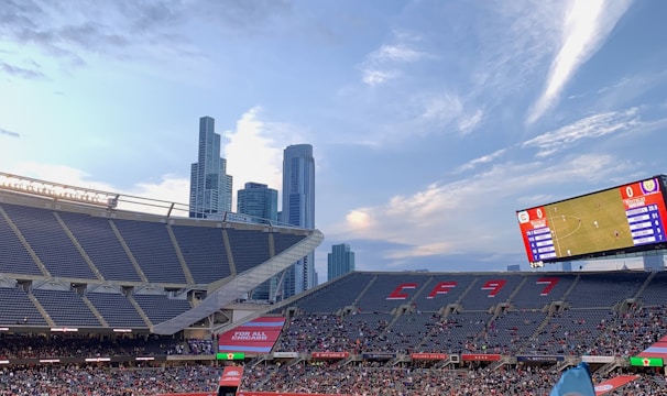 A sports stadium filled with spectators watching a soccer match. Players are on the field in action, and a large scoreboard displays information about the game. The stadium has blue seating and is surrounded by tall city buildings visible in the background against a partly cloudy sky. Fans are dressed casually, some waving flags and wearing team colors.