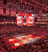 An indoor sports arena filled with spectators and a basketball court illuminated by red lighting. The scoreboards and video screens display promotional content, with numerous banners and championship flags hanging from the ceiling. Players and officials are gathered on the court, possibly in preparation for a game or event.