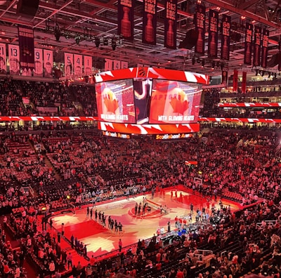An indoor sports arena filled with spectators and a basketball court illuminated by red lighting. The scoreboards and video screens display promotional content, with numerous banners and championship flags hanging from the ceiling. Players and officials are gathered on the court, possibly in preparation for a game or event.