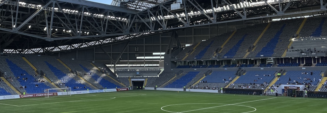 A modern sports stadium with a large, green soccer field and surrounding seating areas. Blue and yellow seats are visible, with some spectators scattered across the stands. The stadium is partially covered with a steel lattice roof, showing a clear blue sky. Several security personnel in high-visibility vests are present.