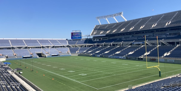 A large, empty sports stadium with blue seats surrounding a well-maintained green grass field. Bright sunlight creates clear shadows, and there are two goal posts on opposite ends of the field. A big electronic scoreboard displays some graphics, and the sky is clear and blue.